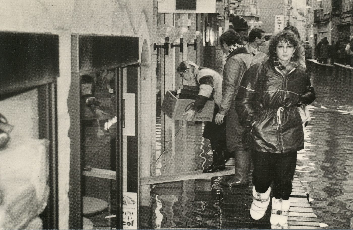 Mâcon. Crue de la Saône. Décembre 1981. Rue Carnot, on évacue ! Les marchandises sont acheminées tant bien que mal vers la place Saint-Pierre, où des stands ont été dressés à la hâte et de nuit. "Que n'a-t-on instauré un sens unique sur ces planches !" semble penser cette cliente qui, par précaution, a chaussé ses bottes.